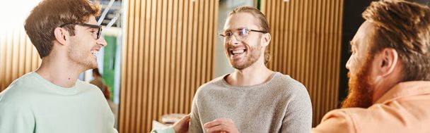 stylish and cheerful businessmen in eyeglasses smiling during discussion of startup project near bearded colleague in modern coworking space, partnership of successful entrepreneurs, banner - Photo, image