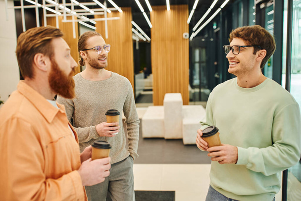 bearded man with stylish and cheerful colleagues in eyeglasses holding takeaway coffee during conversation in lobby of modern coworking space, ambitious businessmen discussing startup project - 写真・画像