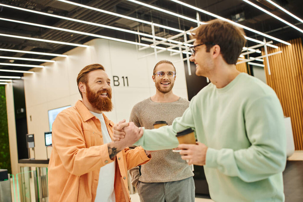 счастливый бородатый мужчина пожимает руку бизнесмену в очках рядом с улыбающимся коллегой, деловые партнеры с кофе идут договариваться в лобби современного коворкинга - Фото, изображение