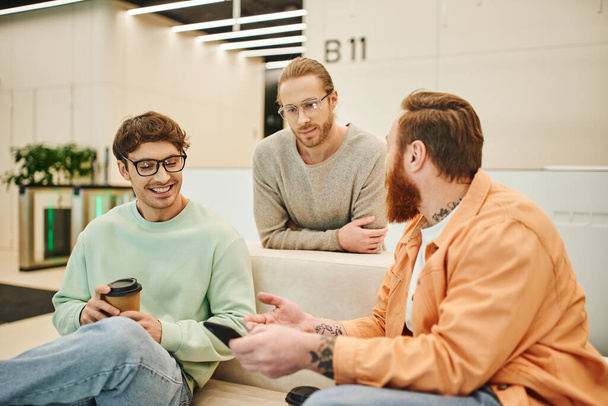 bearded man with mobile phone talking to smiling colleagues in eyeglasses while sitting on comfortable couch during coffee break in lounge of modern office, positive entrepreneurs discussing startup - Fotoğraf, Görsel