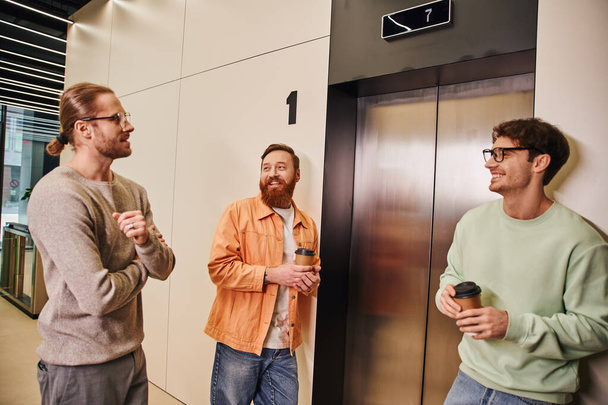 soddisfatti colleghi d'affari in abiti casual alla moda che tengono bevande da asporto in tazze di carta in attesa di ascensore, imprenditori di successo che parlano durante la pausa caffè in ufficio moderno - Foto, immagini