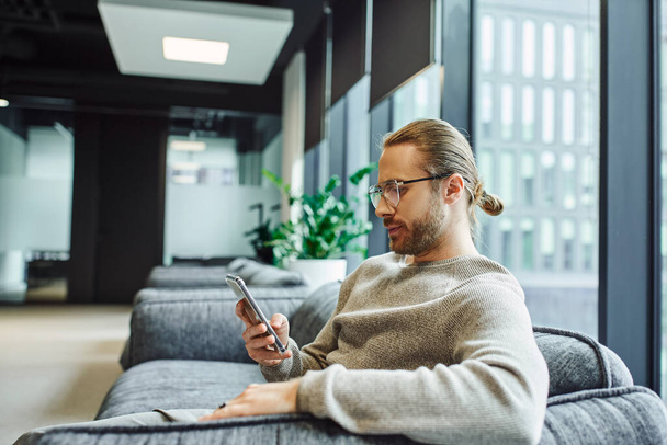 vista lateral de un hombre de negocios serio y elegante en ropa casual y anteojos mirando el teléfono inteligente mientras está sentado en un cómodo sofá en el salón de la oficina contemporánea - Foto, Imagen
