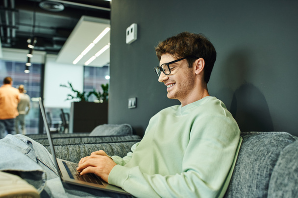 Χαρούμενος επιχειρηματίας με casual ρούχα και κομψά γυαλιά πληκτρολογώντας στο laptop ενώ κάθεται σε άνετο καναπέ στο σύγχρονο γραφείο συνεργασίας, επιχειρηματική έμπνευση και παραγωγικότητα - Φωτογραφία, εικόνα