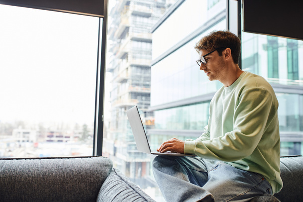 side view of attentive man in casual clothes and eyeglasses sitting near large windows in modern office space and working on startup planning on laptop, business inspiration and ambition concept - Fotoğraf, Görsel