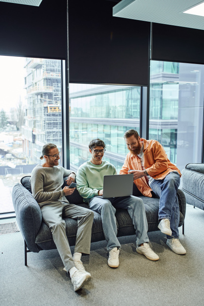 full length of business partners with takeaway drink and mobile phone smiling near colleague working on laptop, sitting on comfortable sofa in lounge of modern coworking office with large windows  - Photo, Image