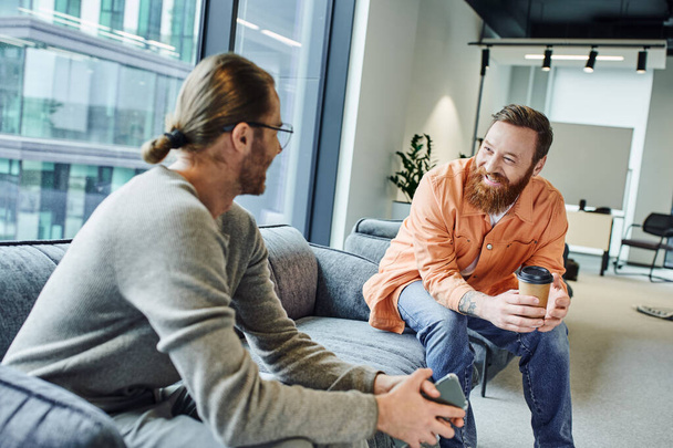 cheerful, bearded and tattooed businessman with coffee to go discussing startup project with colleague sitting with mobile phone on couch in office lounge of modern coworking environment - Photo, Image