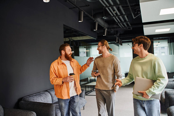 vreugdevolle bebaarde ondernemer houdt koffie te gaan en te praten met zakenpartners met smartphone en laptop wandelen in de hedendaagse lounge van coworking ruimte, succesvolle samenwerking - Foto, afbeelding