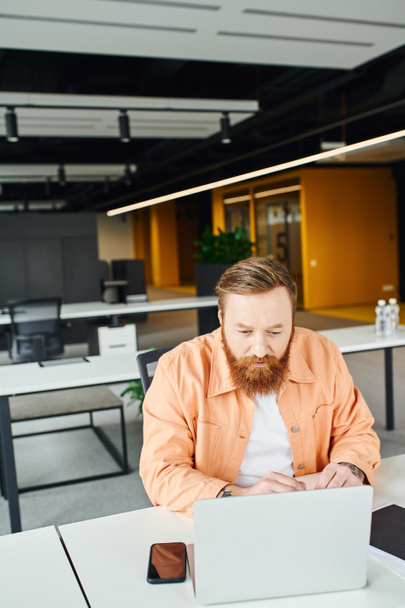 сфокусированный, бородатый и татуированный бизнесмен смотрит на ноутбук, сидя на современном рабочем месте рядом со смартфоном с чистым экраном в современном офисном пространстве и работает над стартап-проектом - Фото, изображение