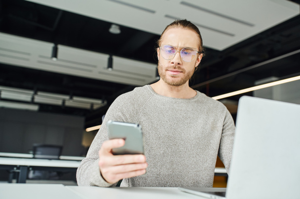 vállalkozó szemüveg és alkalmi ruhák, komoly arckifejezéssel, nézi a mobiltelefon, miközben dolgozik indítási projekt közelében laptop elmosódott előtérben a modern irodahelyiségben - Fotó, kép