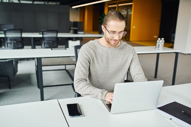 optimistic businessman in casual clothes and eyeglasses planning startup and typing on laptop near folder and smartphone with blank screen at workplace in contemporary coworking office - Photo, Image