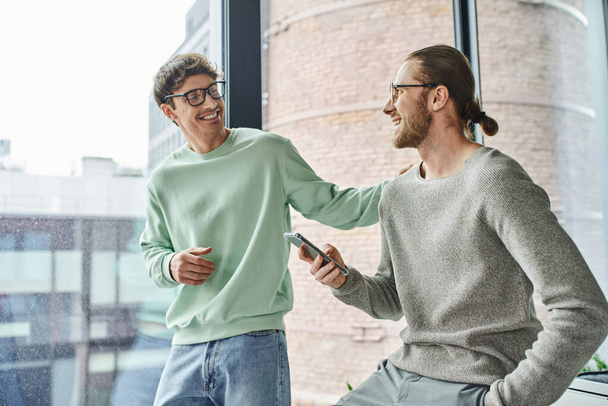 joyful entrepreneur in eyeglasses and casual clothes touching shoulder of business partner sitting with smartphone and hand in pocket near large office window, concept of successful collaboration - Photo, Image