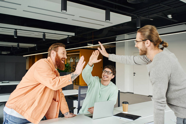 overjoyed bearded and tattooed entrepreneur giving high five to business partners near laptop and takeaway drink in modern environment of coworking office, concept of successful collaboration - Photo, Image