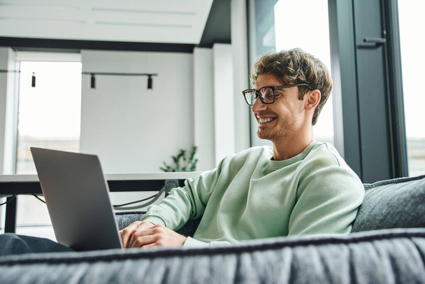 fröhlicher und erfolgreicher Geschäftsmann in stylischer Brille und lässiger Kleidung sitzt auf bequemer Couch in der Bürolounge des Coworking Space und vernetzt sich am Laptop, Geschäftsidee-Konzept - Foto, Bild