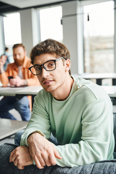 positive and ambitious entrepreneur in eyeglasses and casual clothes looking at camera while his business colleague working on blurred background in contemporary office, productive coworking concept - Photo, Image