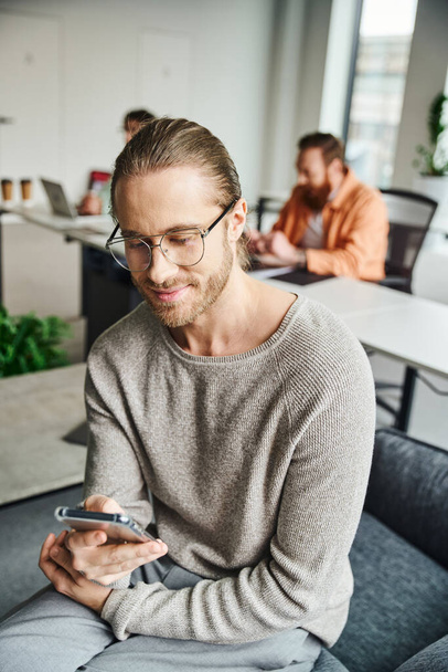 Улыбающийся бизнесмен в очках и повседневной одежде, работающий на смартфоне рядом с коллегами, работающий над размытым фоном, успешной концепцией предпринимательства - Фото, изображение
