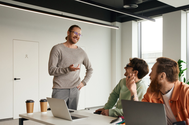 pozitív vállalkozó stílusos szemüveg álló kézzel a csípőn, és ujjal mutogatva, miközben megvitatják az üzleti projekt csapat ül laptopok modern coworking iroda - Fotó, kép