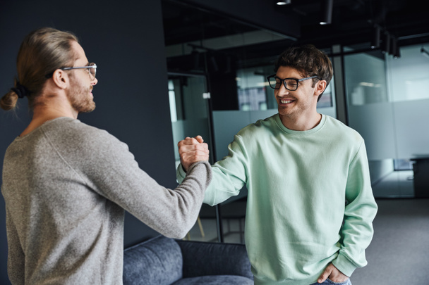 stylish and joyful business partners in eyeglasses and casual clothes confirming agreement and shaking hands in modern lobby of coworking office space, successful collaboration concept - Photo, Image