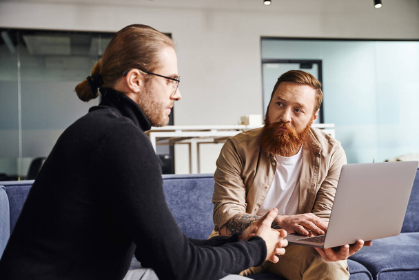 bearded businessman showing startup project on laptop to serious entrepreneur in black turtleneck and eyeglasses while sitting on couch in contemporary office, business partnership concept - Photo, Image