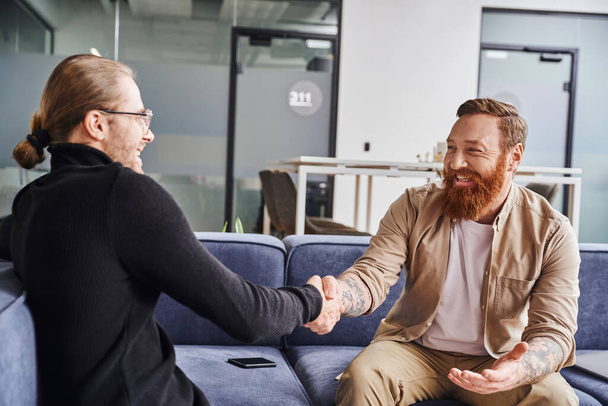 delighted, bearded and tattooed entrepreneur sitting on couch near smartphone and shaking hands with business partner in black turtleneck in contemporary office, successful partnership concept - Photo, Image