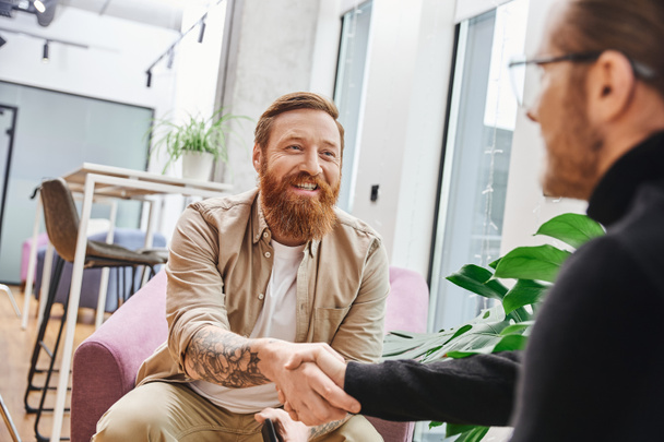overjoyed, tattooed and bearded entrepreneur in casual clothes confirming agreement and shaking hands with businessman on blurred foreground, successful partnership concept - Photo, Image