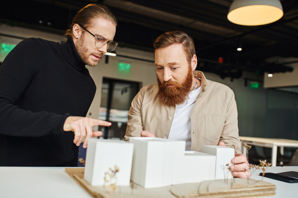 poważny architekt w okularach i czarnym golfie wskazujący na model budynku i rozmawiający z brodatym kolegą pracującym we współczesnym studiu projektowym, architekturze i koncepcji biznesu - Zdjęcie, obraz