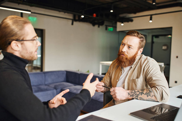 figyelmes, szakállas és tetovált vállalkozó gazdaság toll és hallgat üzleti partner fekete garbó beszél startup projekt közelében laptop kortárs irodai környezetben - Fotó, kép