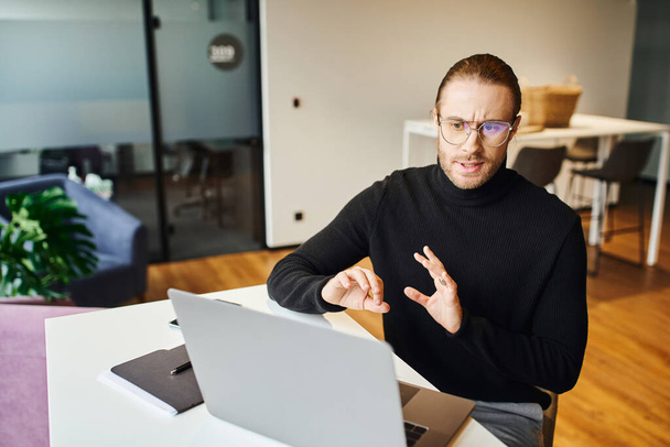 serious entrepreneur in black turtleneck and eyeglasses gesturing and talking while having video call on computer in contemporary office space, business lifestyle concept - Foto, afbeelding