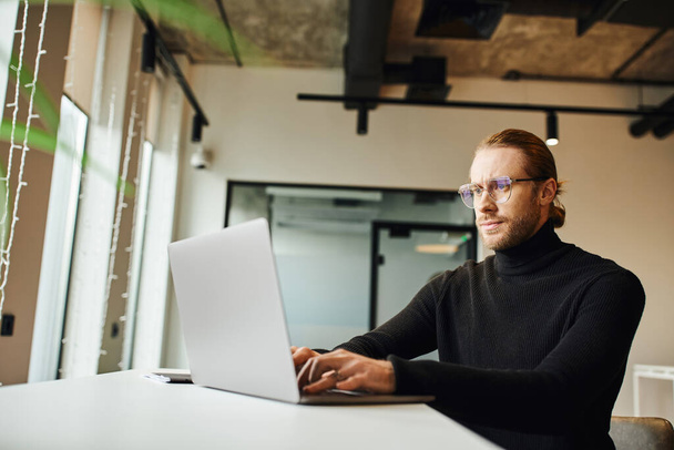 concentrated businessman in black turtleneck and eyeglasses thinking near laptop and working on startup project in modern office environment, business lifestyle concept - Photo, Image