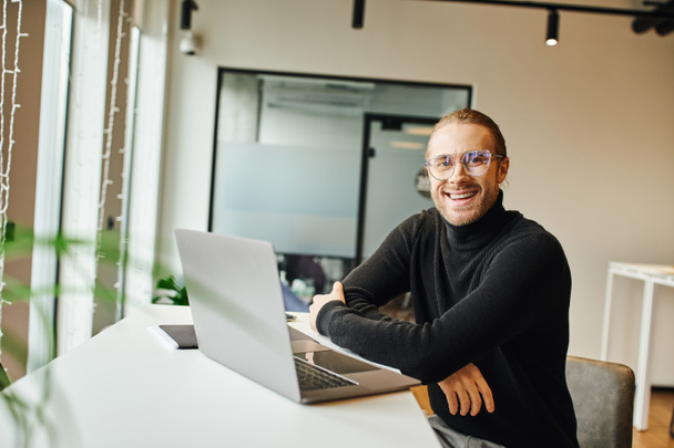 Siyah balıkçı yaka kazak ve gözlüklü neşeli ve şık iş adamı modern ofis ortamında dizüstü bilgisayarın yanında otururken kameraya bakıyor, iş hayatı konsepti - Fotoğraf, Görsel