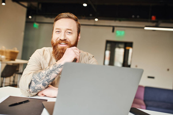 cheerful, bearded and tattooed entrepreneur with radiant smile looking at camera while sitting near laptop and working on startup project, professional headshot, business lifestyle concept - Photo, Image