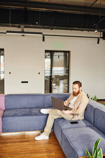 полная длина татуированный и бородатый бизнесмен в стильной повседневной одежде работает на ноутбуке, сидя на удобном диване рядом со смартфоном в гостиной современного офиса, бизнес-концепция образа жизни - Фото, изображение
