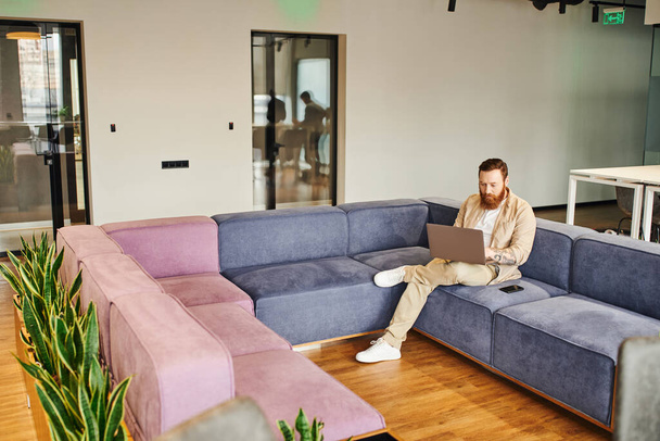 full length of tattooed and bearded entrepreneur sitting on comfortable couch next to mobile phone and working on laptop near green plants in office lounge, business lifestyle concept - Photo, Image