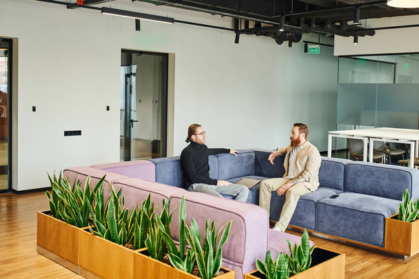 hommes d'affaires sérieux dans des vêtements décontractés élégants assis sur un canapé doux près de plantes vertes et discuter du projet de démarrage dans le salon de bureau moderne avec un intérieur de haute technologie, partenariat et concept de succès - Photo, image
