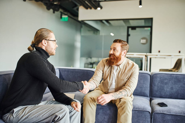 happy bearded and tattooed man closing deal and shaking hands with entrepreneur in black turtleneck in lounge of contemporary coworking office, partnership and success concept - Foto, Imagem