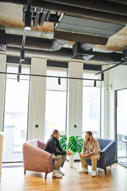 stylish entrepreneurs sitting in cozy armchairs and planning startup project near green plant and large windows in modern office lounge, partnership and success concept - Photo, Image