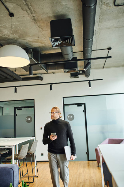 carrière georiënteerde zakenman in zwarte coltrui en stijlvolle bril lopen met laptop en mobiele telefoon, terwijl weg te kijken in de moderne coworking ruimte, succesvolle business concept - Foto, afbeelding