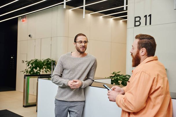 excited businessman in eyeglasses and casual clothes talking to bearded colleague at reception in lobby of contemporary coworking office, entrepreneurs discussing possibility of startup - Photo, Image