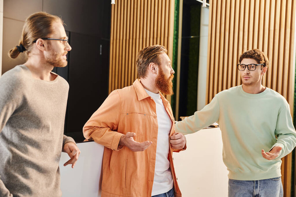 szakállas üzletember komoly szemüveges és alkalmi ruhás kollégákkal beszélget, miközben startup projektet tervez a kortárs coworking környezet halljában, üzleti együttműködést - Fotó, kép
