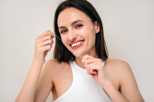 studio shot of smiling woman holding dental floss in hands, looking at camera, cleaning teeth, standing on beige copy space background. healthy oral care habit for caries prevention - Photo, Image