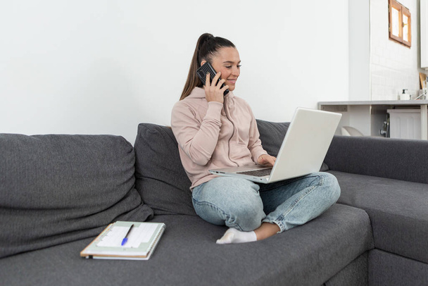 Hymyilevä nainen istuu sohvalla lootusasennossa, puhuu puhelimessa tehdessään ostoksia verkossa kannettavan tietokoneen kanssa kotoa. - Valokuva, kuva