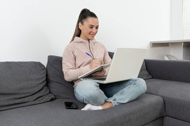 Günlük kıyafetlerle gülümseyen genç bir kadın evdeki kanepede oturuyor dizüstü bilgisayarını tutarken bir defter yazıyor. - Fotoğraf, Görsel