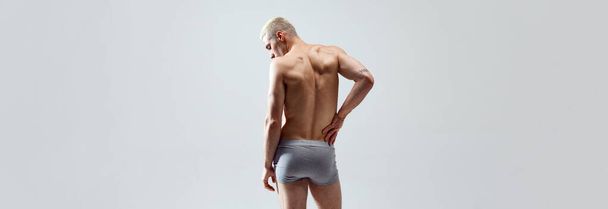 Rear view image of young man with muscular, relief, fit, strong healthy body, back, posing in underwear shirtless against grey studio background. Concept of male beauty, body care, health, sport, ad - 写真・画像