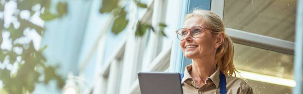 Middle aged caucasian business woman with tablet looking sideways and smiling while standing near garden house. Small business concept - Photo, Image