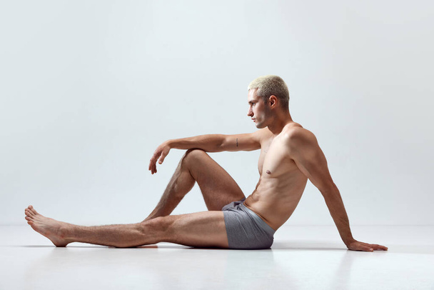 Young handsome man with muscular, relief, fit, strong body posing on floor in underwear against grey studio background. Concept of male natural beauty, body care, health, sport, fashion, ad - Valokuva, kuva