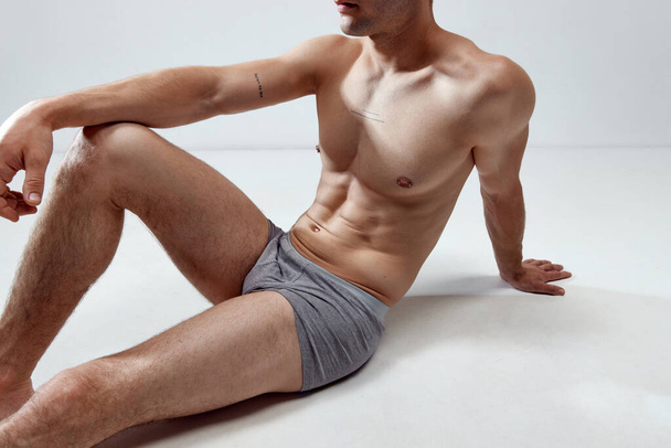Cropped image of young man with muscular, relief, fit, strong body posing on floor in underwear against grey studio background. Concept of male natural beauty, body care, health, sport, fashion, ad - 写真・画像