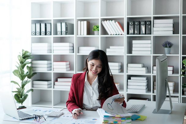 Красивая азиатка-бизнесвумен, работающая на ноутбуке и рассчитывающая с помощью финансового калькулятора из статистических графиков, графиков. Успешные результаты бизнеса в современном офисе носят красную рубашку. Высокое качество - Фото, изображение