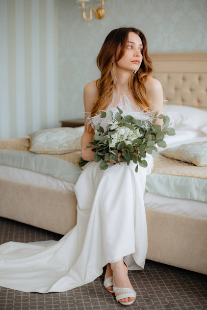 Amazing young bride wearing wedding dress adorned with fluffy feathers, white high heels shoes sitting on bed with big bouquet of blossoms in hands. Charming lady with brown hair looking aside. - Фото, изображение
