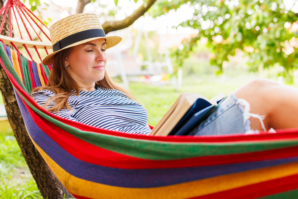 Молодая женщина читает книгу, лежа в удобном гамаке в зеленом саду. - Фото, изображение