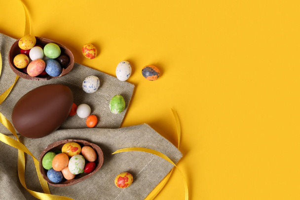 Whole and halves of chocolate eggs with colorful candies on yellow background, flat lay. Space for text - Photo, Image