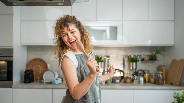 One happy young adult caucasian woman wear apron in the kitchen smile hold mixing spoon sing - Foto, afbeelding
